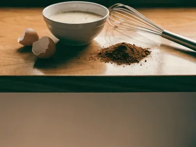 A bowl of cake batter with a whisk, cocoa powder and eggshells on a wooden counter