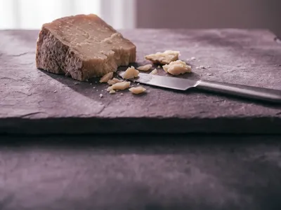 A wedge of aged hard cheese with crumbles and a knife on a plum-grey stone slab