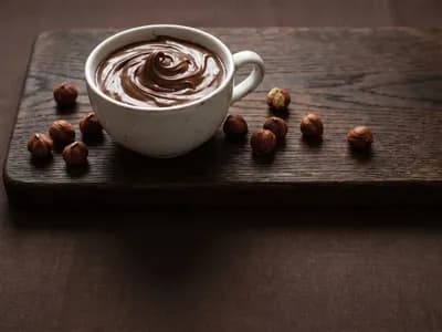 A swirl of rich chocolate spread in a ceramic cup with toasted hazelnuts on a dark wood board