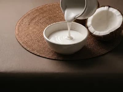 Coconut cream pouring from a jug into a bowl beside a fresh halved coconut