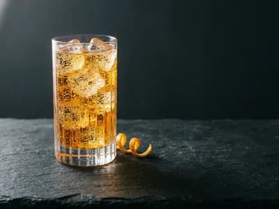 An effervescent amber drink with ice cubes and citrus twist on a dark stone surface