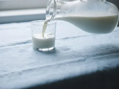 Fresh milk pouring from a glass carafe into a tumbler on a mist-blue cloth
