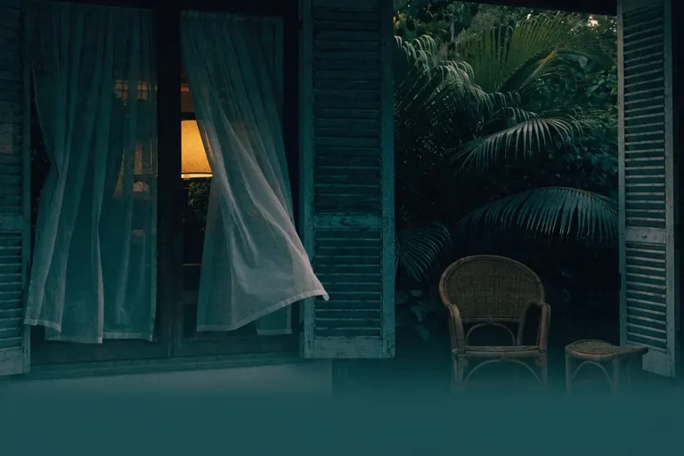 A tropical verandah at dusk with rattan furniture, louvered shutters and sheer curtains drifting in the breeze