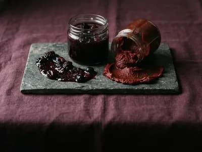 Open jars of dark fruit preserve and thick tomato paste with a smear of each on a stone slab