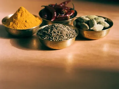Brass bowls of turmeric, red chilli powder, cumin seeds and green cardamom on a terracotta surface