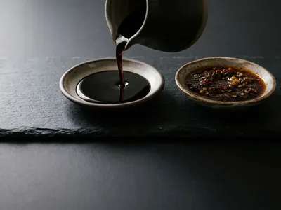 A dark glossy sauce pouring from a ceramic jug into a shallow dish beside chilli-garlic sauce on black slate