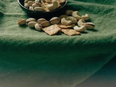 Shelled pistachios, cashews and savoury crackers spilling from a bowl onto a deep olive-green linen cloth