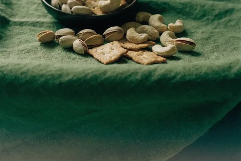 Shelled pistachios, cashews and savoury crackers spilling from a bowl onto a deep olive-green linen cloth