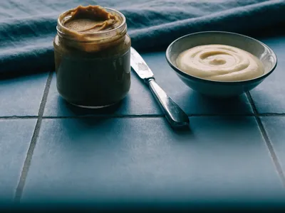An open jar of peanut butter and a bowl of mayonnaise with a butter knife on a slate-blue tiled surface