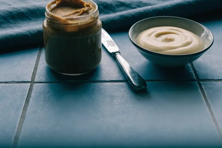 An open jar of peanut butter and a bowl of mayonnaise with a butter knife on a slate-blue tiled surface