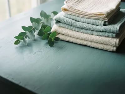 A stack of folded soft cotton wipes beside a eucalyptus sprig on a blue-green surface