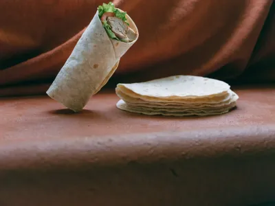 A filled chicken wrap sliced to show grilled chicken, lettuce and tomato, beside a stack of soft flour tortillas