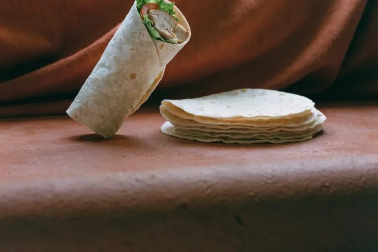 A filled chicken wrap sliced to show grilled chicken, lettuce and tomato, beside a stack of soft flour tortillas