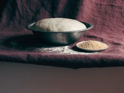 A proving bowl of risen bread dough beside a dish of dried yeast granules and scattered flour on a plum-burgundy cloth