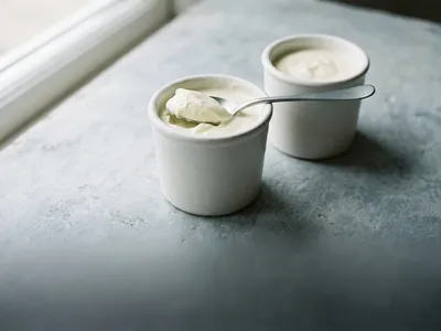 Thick set yogurt in a white ceramic pot with a spoon lifting a curl from the surface