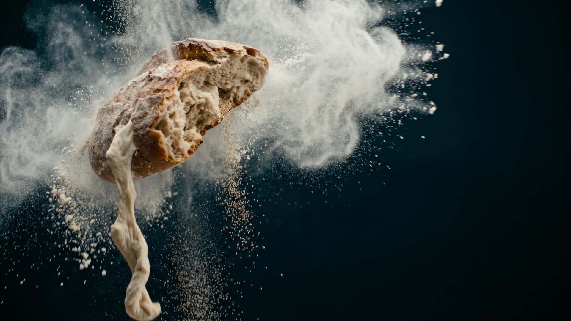 A torn loaf of artisan bread with an open, airy crumb suspended inside a billowing cloud of fine white flour