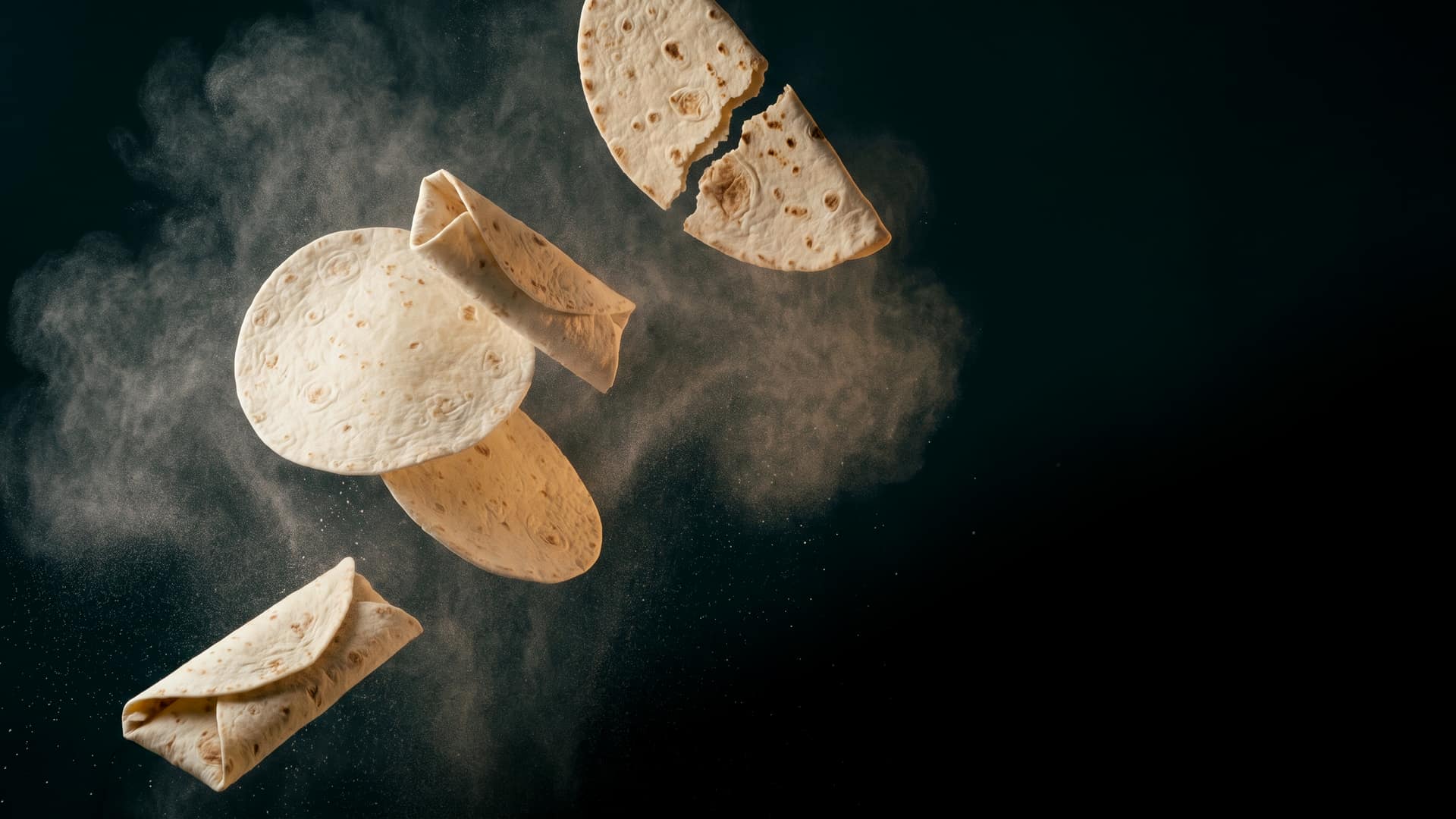 Soft round flour tortillas and wraps suspended weightlessly in mid-air, one folded and one loosely rolled, with a thin crispy piadina cracked in half above them and a fine haze of wheat flour drifting through
