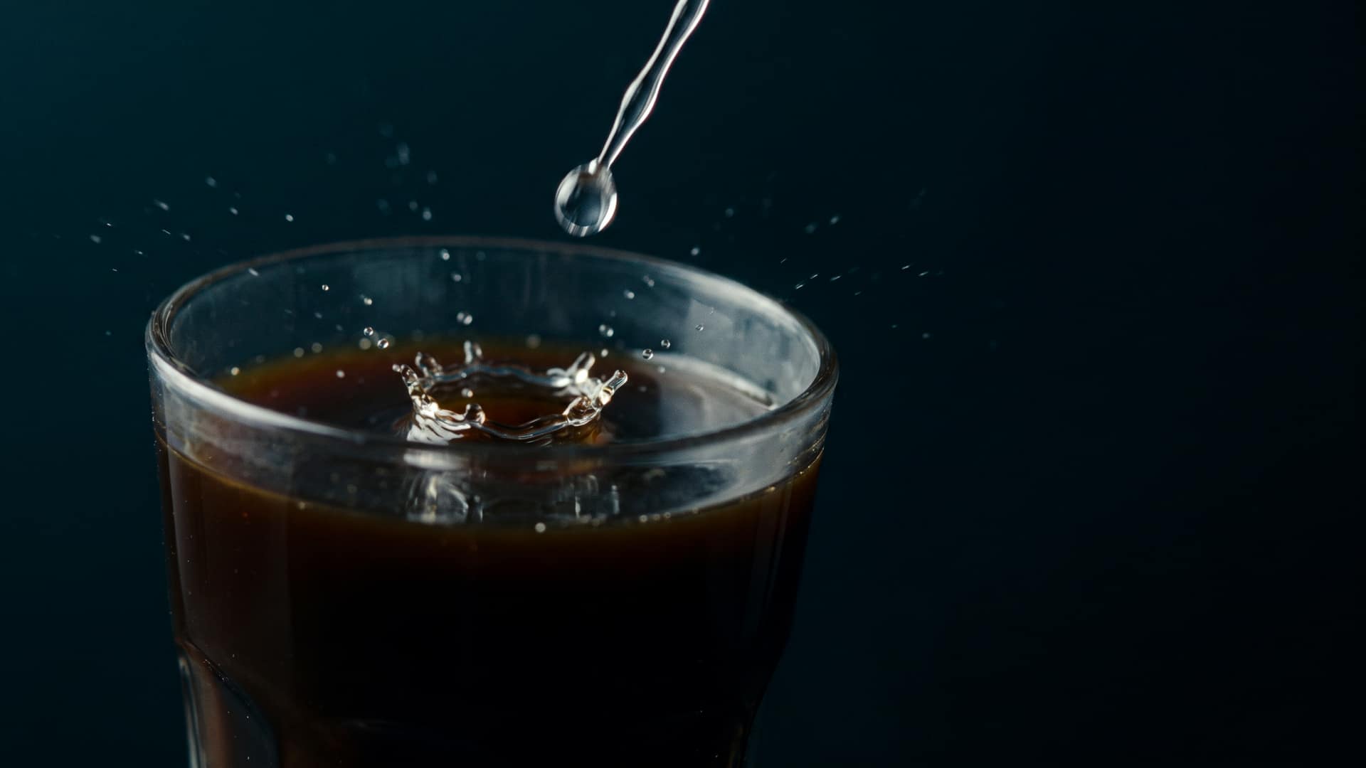 A single clear droplet of liquid sweetener falling into a glass of dark espresso, frozen at the instant of impact
