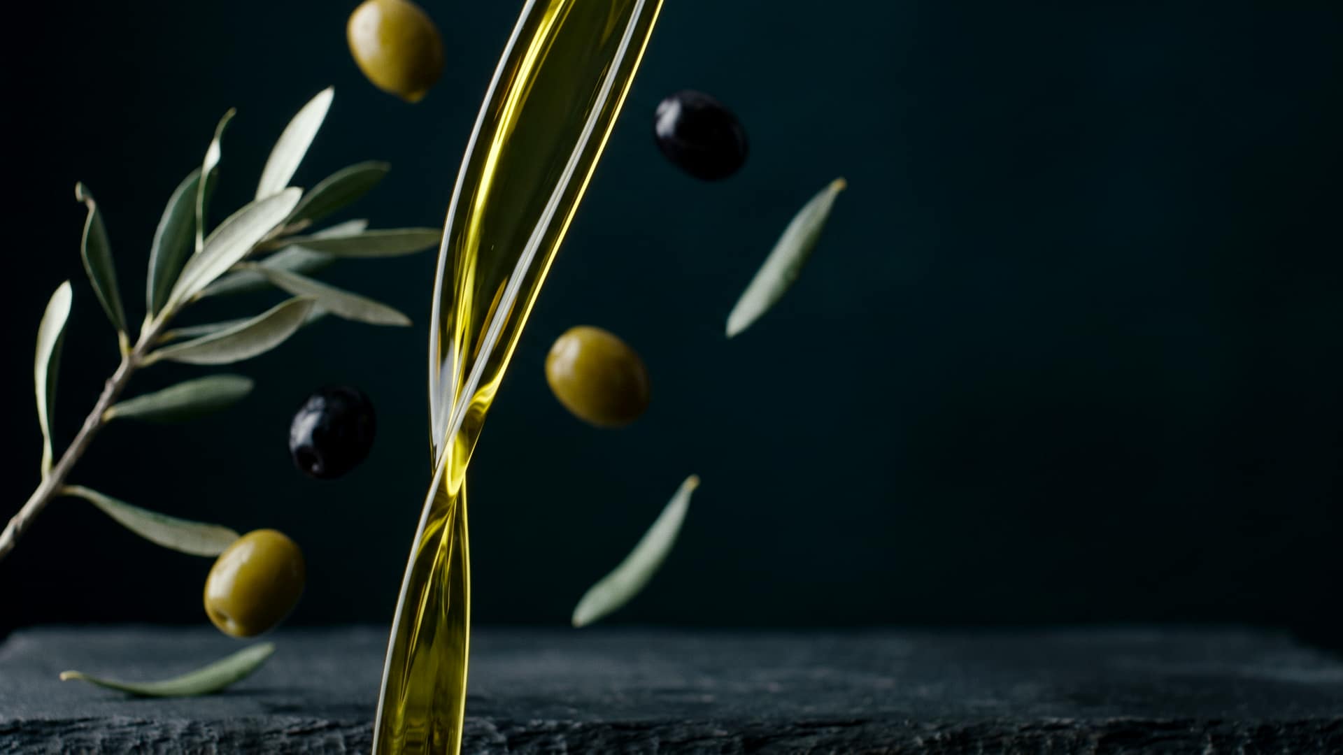 A ribbon of green-gold extra virgin olive oil pouring in mid-air with whole green and black olives and an olive branch suspended around it