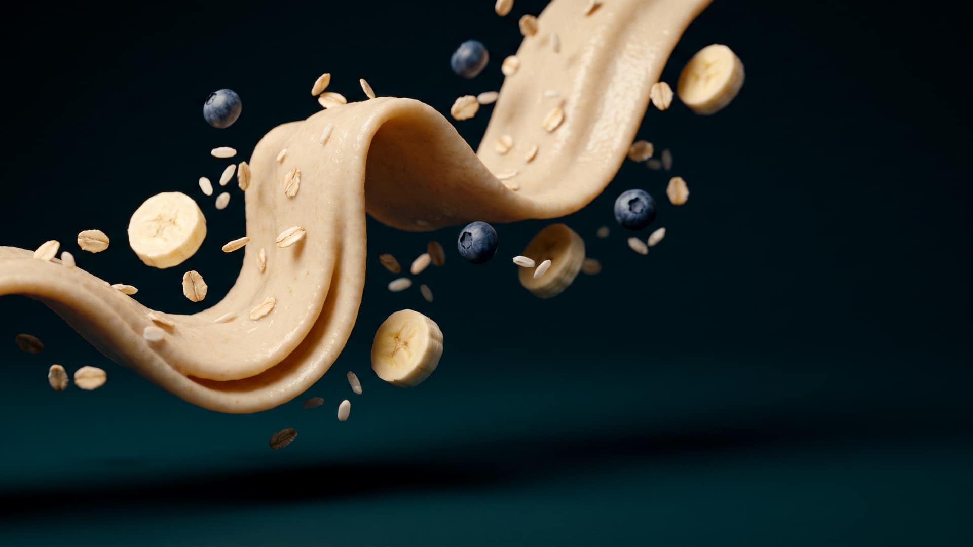 A gentle ribbon of warm milky infant cereal folding through the air with soft oat flakes, banana slices and blueberries suspended around it