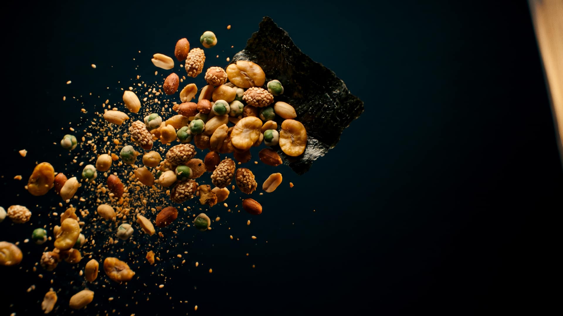 Roasted peanuts, sesame-coated peanuts, wasabi green peas and crisp broad beans scattering outward through a dust of toasted sesame, with a sheet of dark nori curling behind