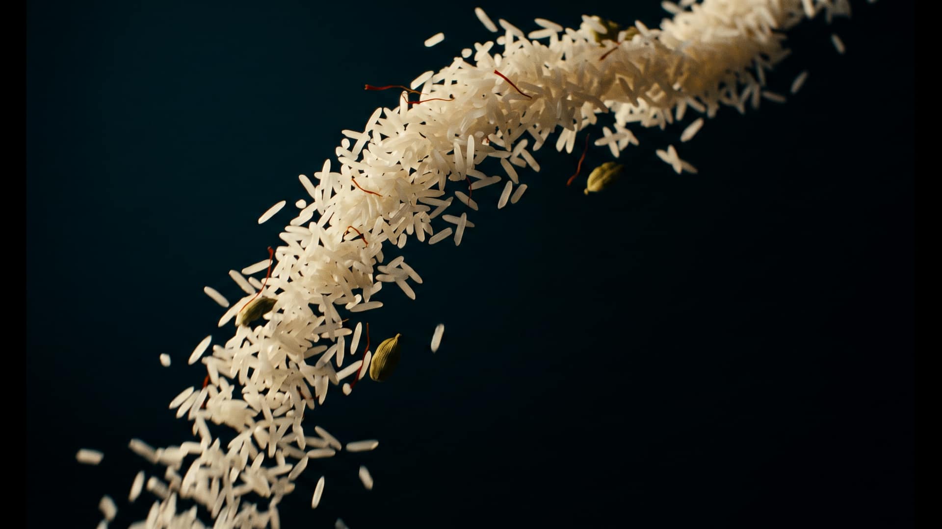 A cascading arc of long white basmati rice grains catching the light, with saffron threads and green cardamom pods drifting among them