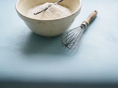 A ceramic mixing bowl with sifted cake flour and a wire whisk on a powder blue surface