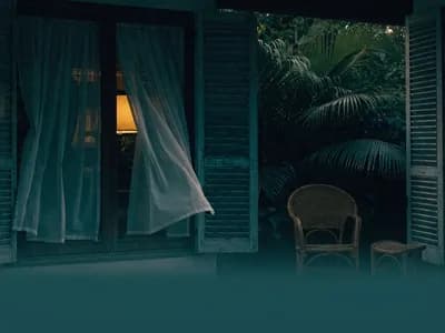 A tropical verandah at dusk with rattan furniture, louvered shutters and sheer curtains drifting in the breeze