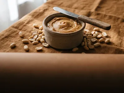 Creamy peanut butter in a stoneware pot with roasted peanuts alongside on a tan cloth