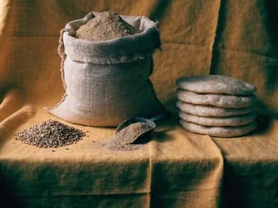 An open sack of wholemeal atta flour with a scoop, loose grain and stacked flatbread on a mustard-yellow cloth
